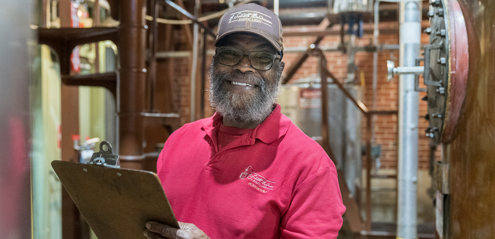 Man in a red polo shirt and blue hat holding a clipboard in a distillery.