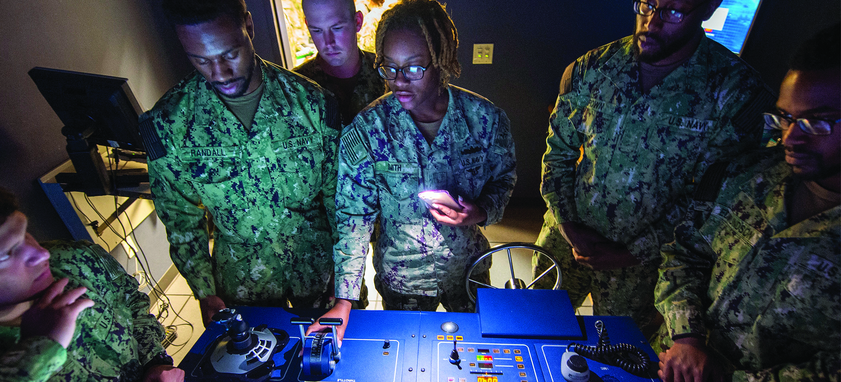 Members of the U.S. Navy gather around a table with equipment embedded into the top