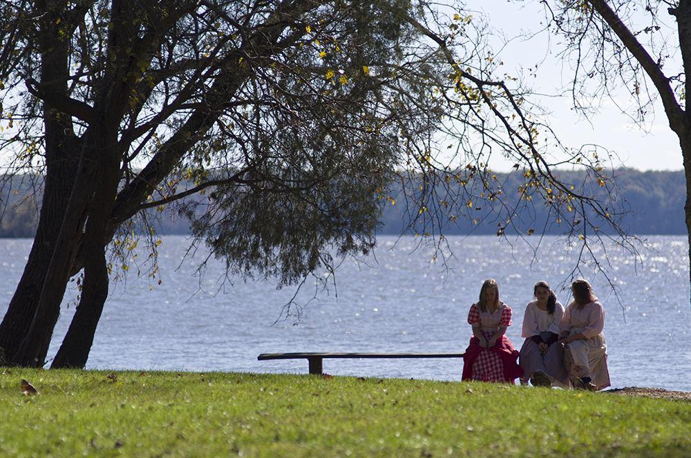 Three women in period clothing sitting on a bench by a lake