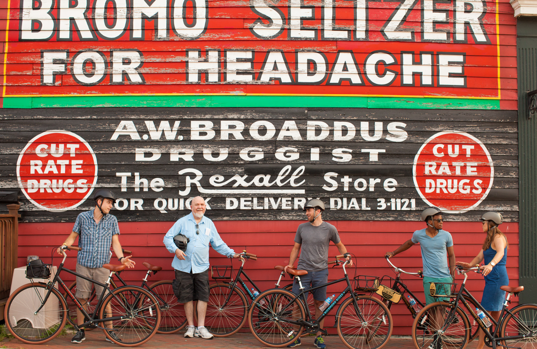 Cyclists pose with their bikes in front of a vintage drugstore mural